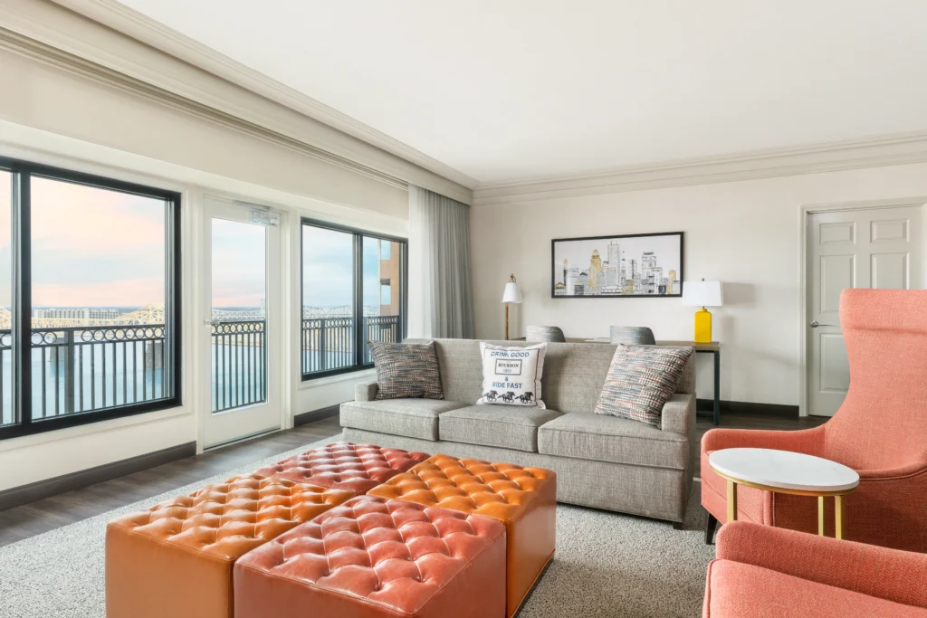 A luxurious living room featuring a gray sofa, two orange leather ottomans, and decorative cushions. Large windows display a city skyline at sunset. Elegant interior design elements enhance the cozy atmosphere.