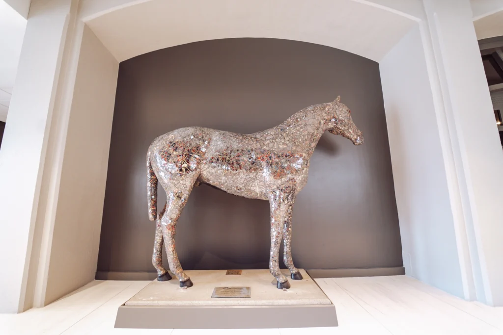 A sculptured horse statue, encrusted with various small, colorful objects, displayed in an arched gray alcove on a white platform.