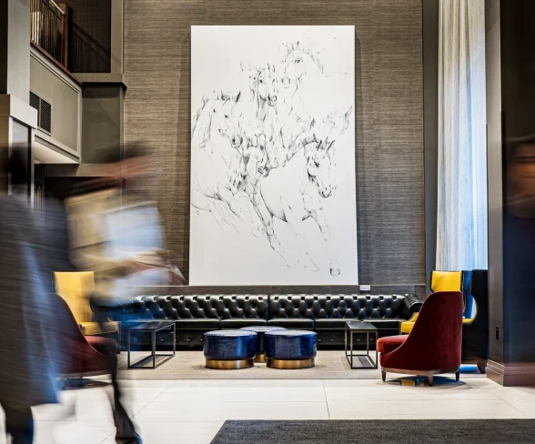 Elegant Lobby with Large Horse Artwork and Passing People A modern hotel lobby featuring a large white canvas with a sketch of galloping horses, flanked by blurred figures in motion, a black tufted sofa, and colorful chairs.